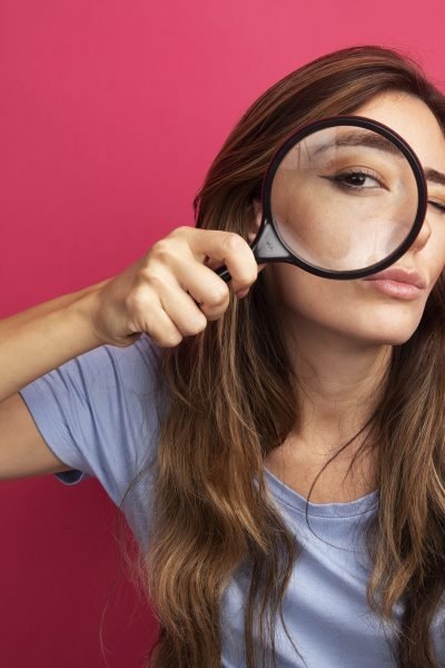 young beautiful woman in blue t-shirt looking at camera through magnifying glass with interest standing over pink background